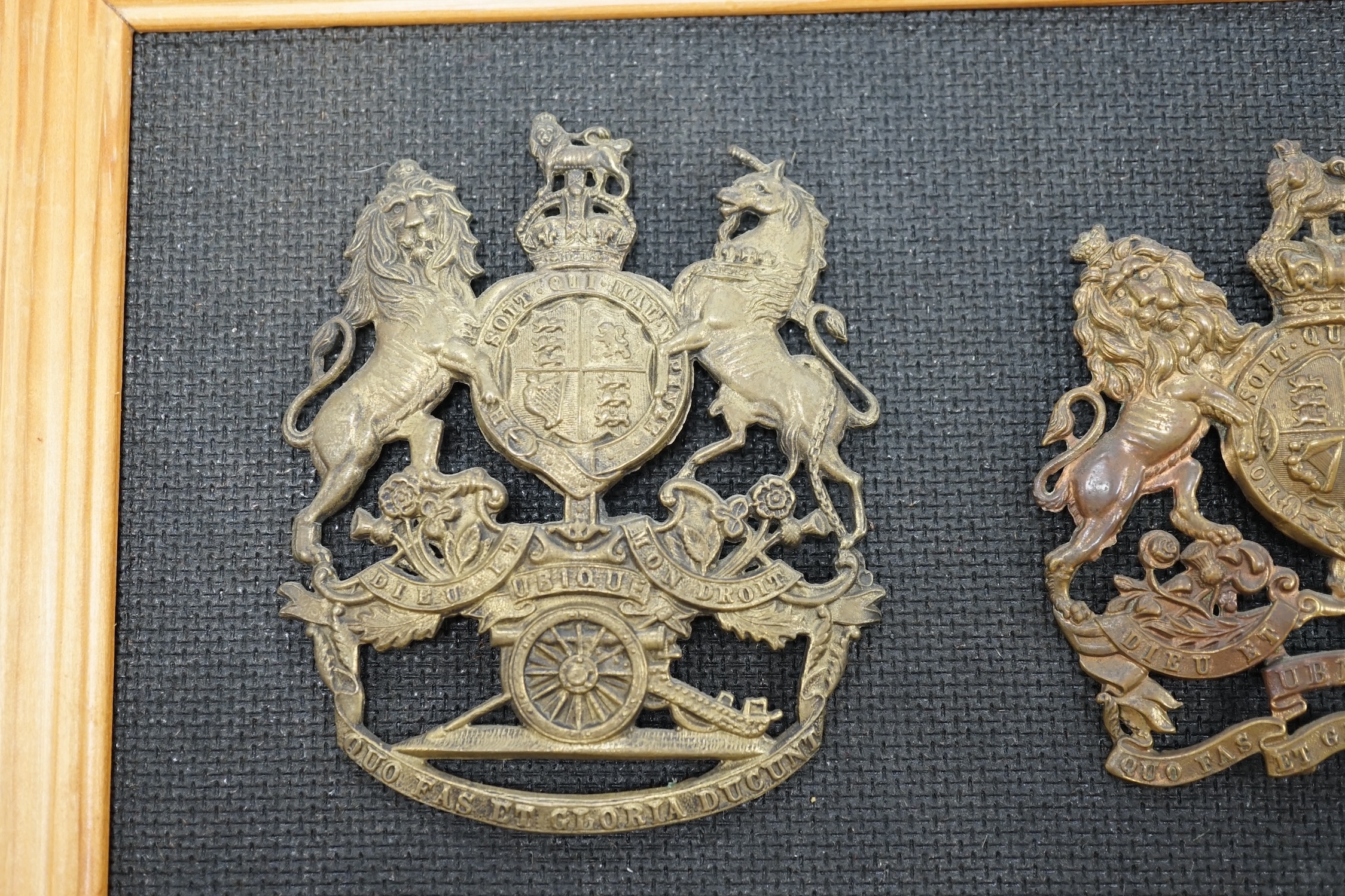 Two Royal Artillery brass helmet plates, one with three lugs and the other with two lugs, loosely mounted onto a backing board. Condition fair to good, top lug missing to one